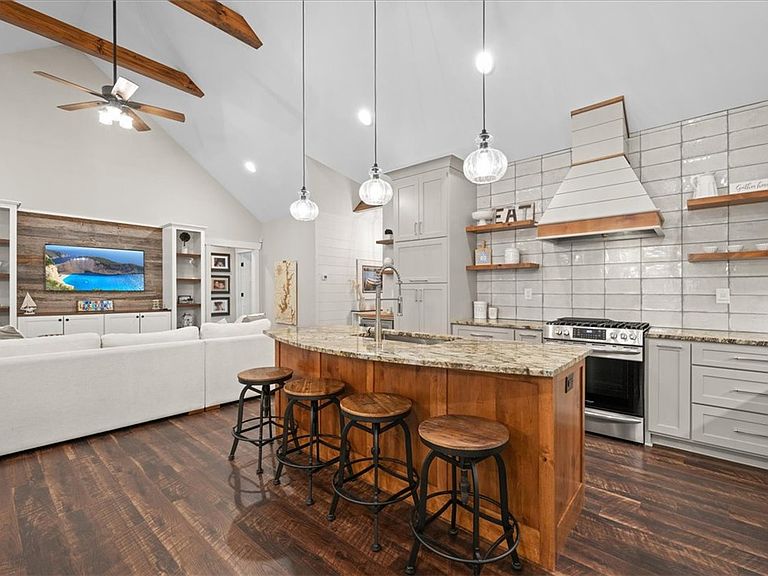 Open-concept great room and kitchen at 107 Cedar Bluff Ct, featuring a vaulted ceiling with stained wood beams, a custom wood range vent hood, a butcher-block island, and four iron-and-wood counter stools